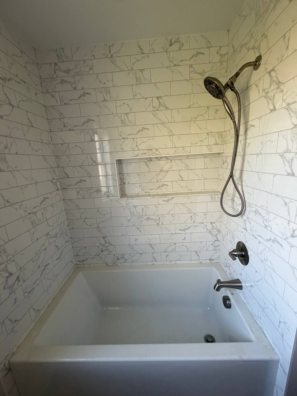 Tiled tub and shower alcove with marble-look subway tile, niche, and handheld showerhead